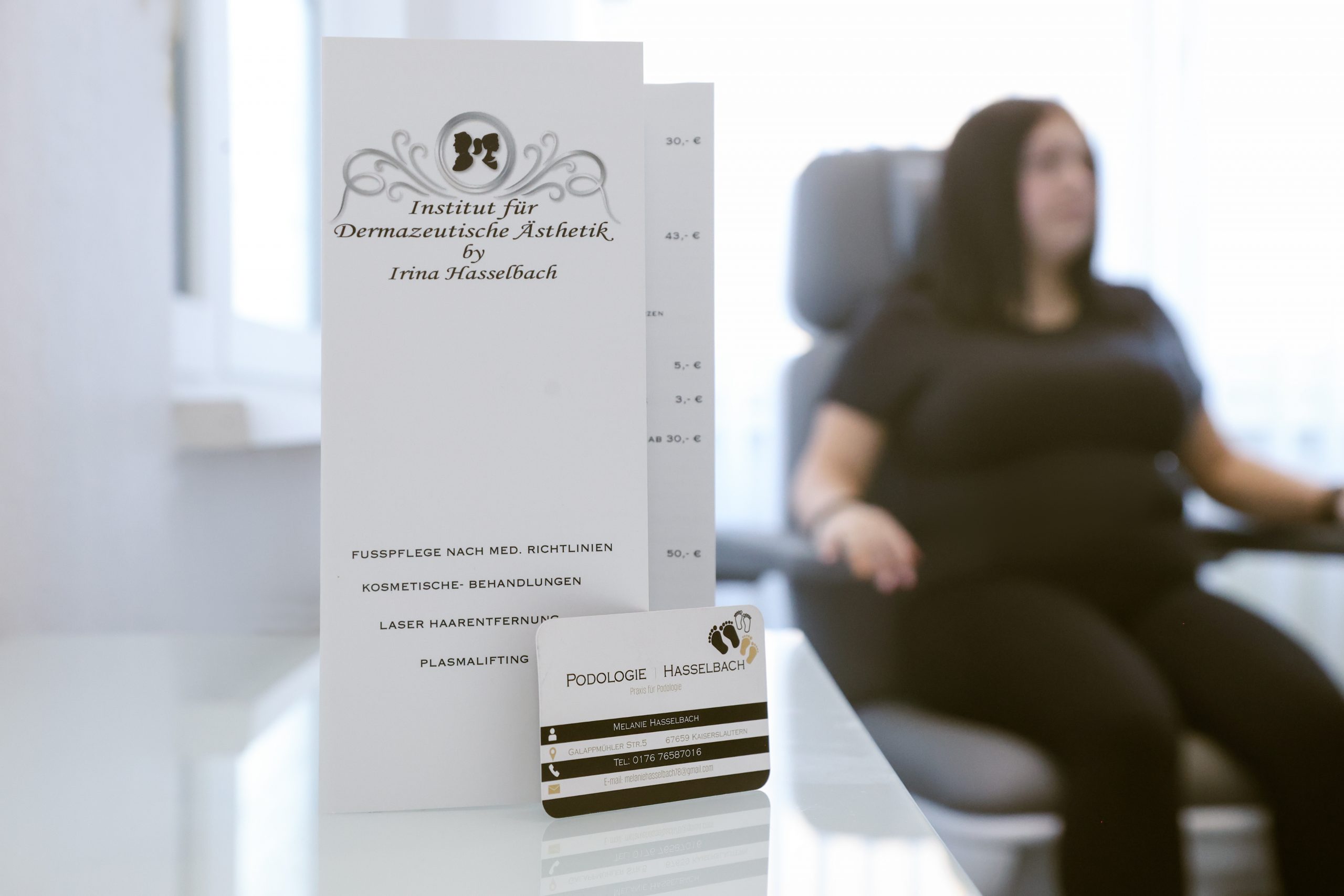 Podologe Kaiserslautern – Praxisfolder und Visitenkarte von Podologie Hasselbach auf einem Tisch, im Hintergrund eine Patientin im Behandlungsstuhl. Kosmetikteam Kaiserslautern. Teamfoto im Kosmetikstudio Kaiserslautern – Irina und Melanie Hasselbach gemeinsam im Behandlungsraum